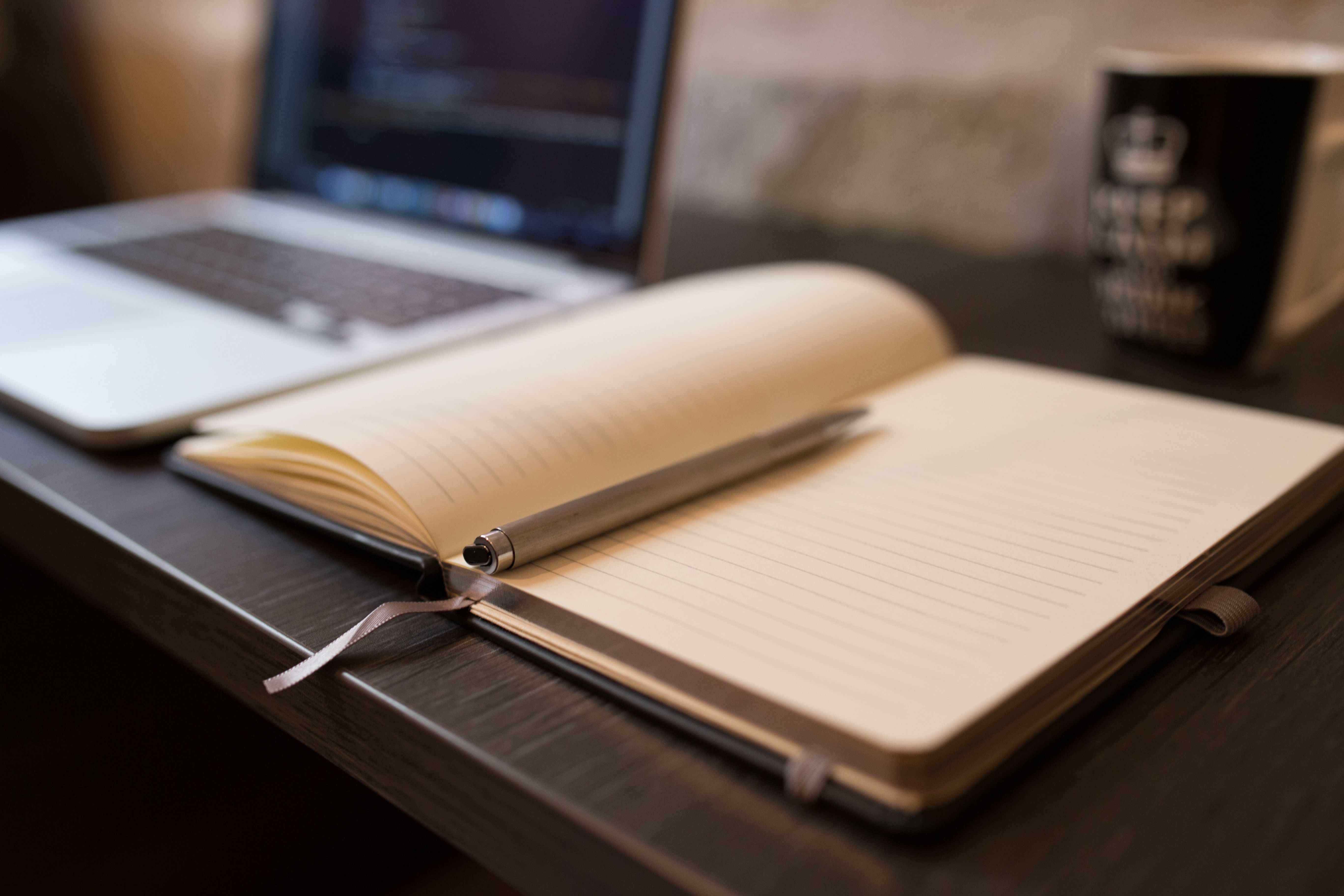 An open blank notebook with a pen resting on it, beside a laptop and a mug on a dark wood desk, warm indoor lighting.